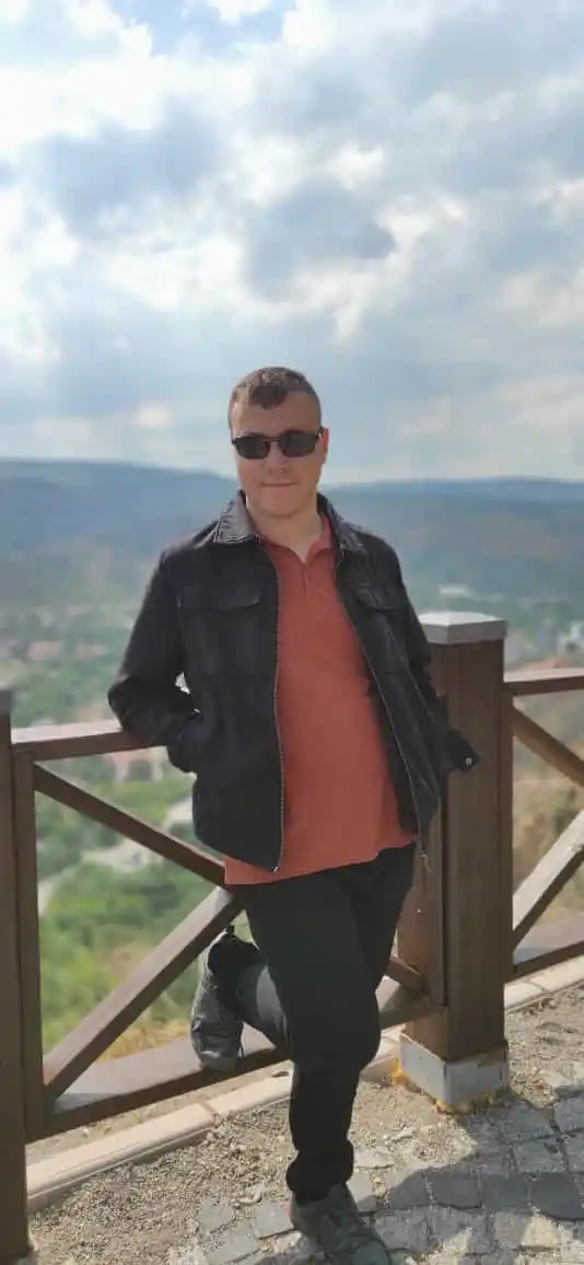 Muhammet Enes Şenovalı, arka planda geniş bir vadi ve dağ manzarasının uzandığı açık bir seyir alanında, ahşap bir korkuluğa sağ koluyla yaslanmış halde ayakta duruyor. Üzerinde siyah bir ceket, somon rengi yakalı bir tişört, siyah pantolon ve koyu renkli spor ayakkabılar var. Ayrıca siyah güneş gözlüğü takıyor. Kısa kesilmiş kahverengi saçlara ve sakalsız, net bir yüze sahip. Kameraya doğru, elleri cebinde ve sol bacağını arkaya doğru kıvırıp korkuluğa dayamış rahat bir duruşla, hafif bir tebessümle bakıyor. Arka plandaki yeşil tepeler, uzaklardaki yerleşim yeri ve gökyüzündeki parçalı bulutlu aydınlık hava, fotoğrafa huzurlu ve rahat bir doğa gezisi atmosferi katıyor.
